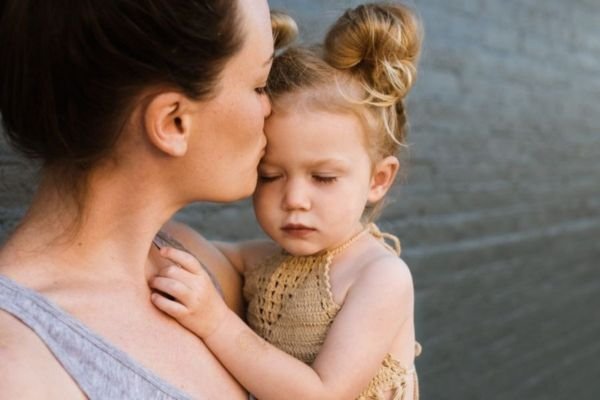 A woman holds a little girl close, kissing her on the cheek in a display of love and care